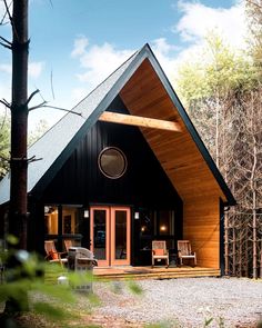 a small black cabin in the woods with two chairs on the front porch and one door open