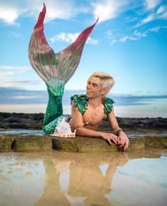 a woman with blonde hair and green dress laying on the ground next to rocks near water