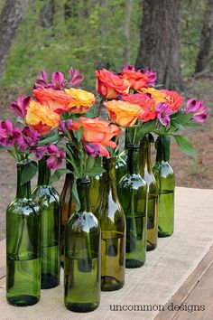 several green glass bottles with flowers in them on a wooden table near grass and trees