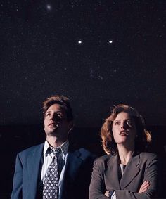 two people standing in front of a night sky with the stars above them and one person wearing a suit