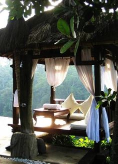 a gazebo with white curtains and pillows on the floor next to trees in front of it