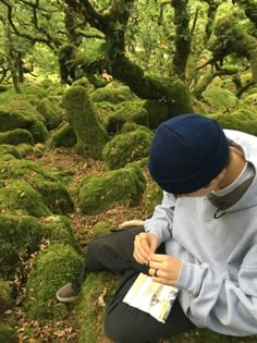 a young man sitting on the ground in front of moss covered rocks with trees behind him