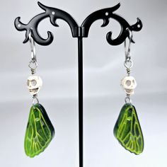 a pair of green and white earrings on a black metal stand with a skull hanging from it's ear