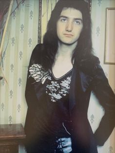 a man with long black hair standing in front of a wallpapered background and looking at the camera