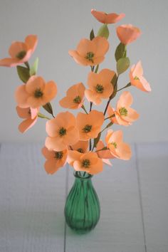 a green vase filled with pink flowers on top of a wooden table next to a white wall