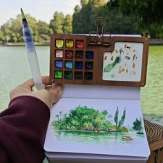 a person holding an art book with watercolors on it and a pen in their hand