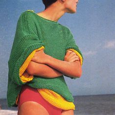 a woman standing on the beach with her arms crossed wearing a green and yellow sweater