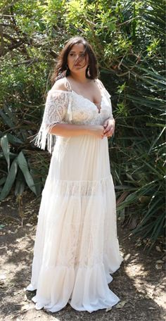 a woman wearing a white dress standing in front of some bushes and trees with her arms crossed