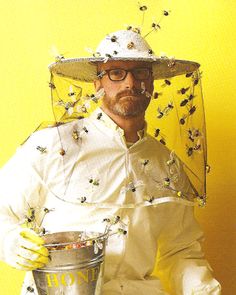 a man dressed in bee suits and holding a bucket with bees around his neck, standing against a yellow background