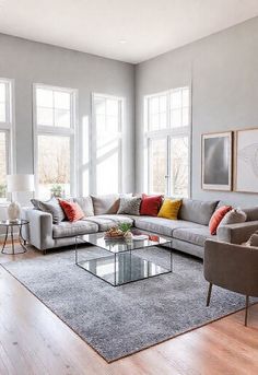 A spacious, contemporary living room featuring soft grey walls, a plush grey sofa with colorful throw pillows, a sleek glass coffee table, and minimalistic art pieces. The space is well-lit with natural sunlight streaming through large windows, complemented by neutral wood flooring and a cozy grey area rug. Modern Grey Living Room, Grey Sofa, Minimalistic Art, Colorful Throw Pillows, Well Lights, Gray Sofa, Natural Sunlight