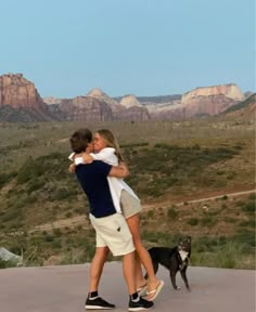 two people hugging each other with a dog in the foreground and mountains in the background