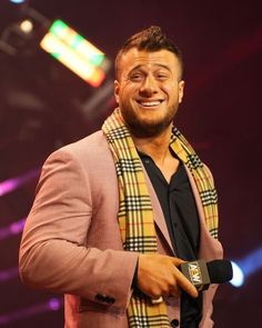 a man in a pink suit and scarf smiling at the camera while holding a microphone