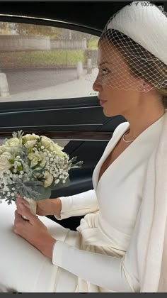 a woman in a white dress and veil sitting in a car holding a bouquet of flowers