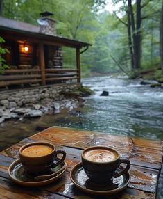 two cups of coffee sitting on top of a wooden table next to a river in the woods