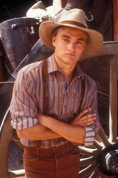 a young man wearing a hat and suspenders standing in front of a horse drawn carriage