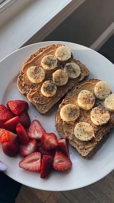 two pieces of toast with bananas and strawberries on a white plate next to a window