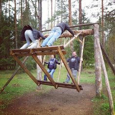 three people are playing on a wooden swing set in the woods while another person is standing next to them