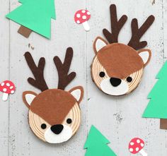 two felt deer head brooches with mushrooms and trees in the background on a white wooden surface