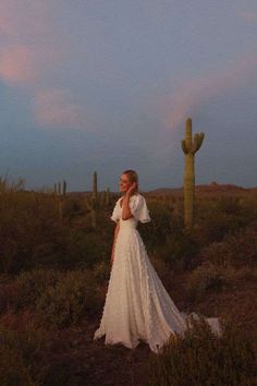 a woman in a white dress standing next to a large green cactus at dusk with the sun setting behind her