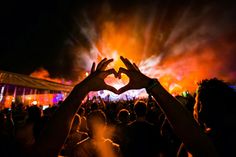 two hands in the shape of a heart over a crowd at a music festival with bright lights