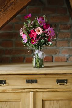 a vase filled with pink and purple flowers sitting on top of a wooden dresser next to a brick wall