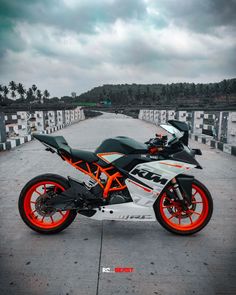 an orange and black motorcycle is parked on the concrete near a bridge with palm trees in the background