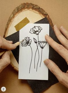 two hands holding a piece of paper with flowers drawn on it, next to a slice of wood