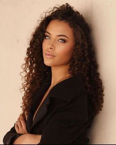 a woman with long curly hair leaning against a wall and looking at the camera while wearing a black shirt