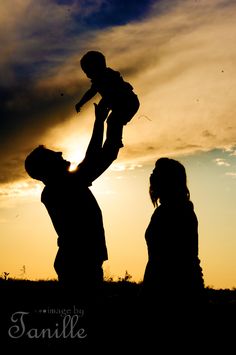 a man holding a child up in the air while standing next to a woman at sunset