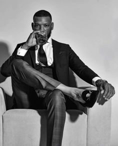 a man in a suit and tie sitting on a chair drinking from a glass with his feet up