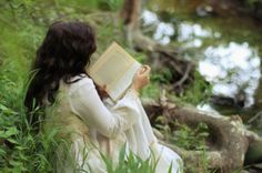 a woman sitting on the ground reading a book next to a river and trees in the background