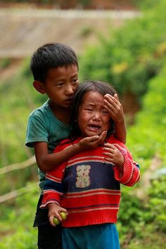 two young children are hugging each other in the grass and dirt, with trees behind them