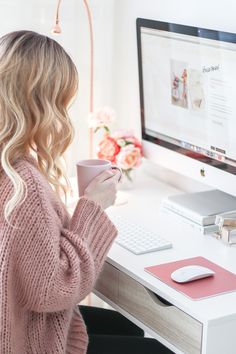 a woman sitting in front of a computer holding a cup and looking at the screen