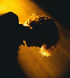 a man with long hair standing in front of a yellow light and holding his head to the side