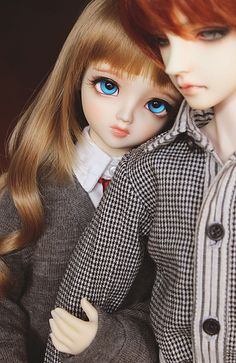 two dolls are posed next to each other, one is wearing a suit and the other has blue eyes