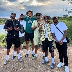 four men standing together in front of a body of water with trees and clouds behind them