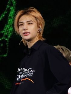 a man with blonde hair wearing a black shirt and earring stands in front of a green screen