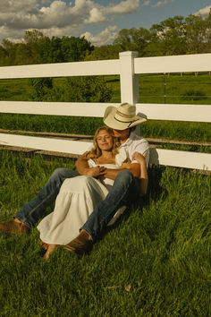 a man and woman sitting on the grass in front of a white fence with a green field behind them