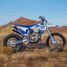 a blue and white dirt bike parked on top of a dry grass covered field with mountains in the background