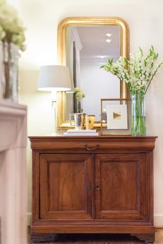 a wooden cabinet sitting in front of a mirror and vase with flowers on top of it