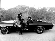 two men sitting on the back of a car with another man standing next to it