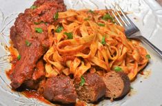 a white plate topped with pasta and meat next to a fork on top of a table