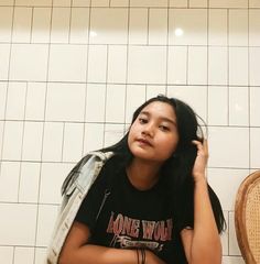 a woman sitting on the floor in front of a white tiled wall with her hand under her hair