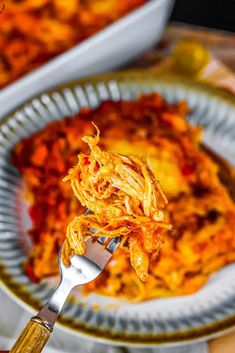 a person holding a fork full of food on a plate with another dish in the background