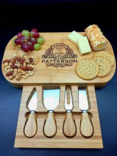 a wooden cutting board with cheese, crackers and grapes on it next to utensils