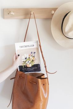 a person holding a book in their hand next to a purse and hat on the wall