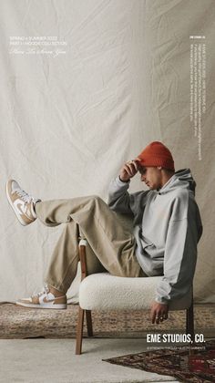 a man sitting on top of a white chair in front of a white wall wearing an orange hat