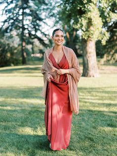 a woman in a long red dress standing on the grass with trees in the background