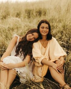 two women are sitting in the tall grass and one is holding her arm around another woman's shoulder