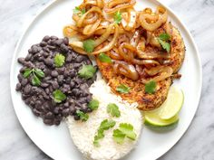 a white plate topped with rice, beans and meat covered in onions next to lime wedges